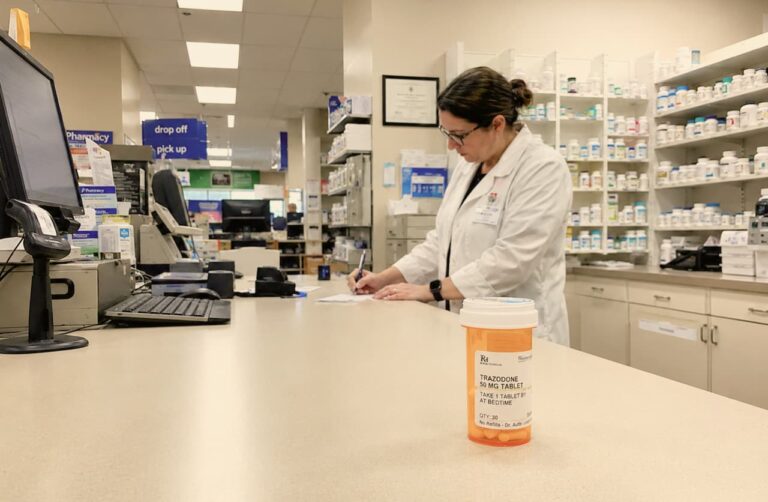 Pharmacist writing at counter with a trazodone 50mg prescription bottle in foreground at a retail pharmacy