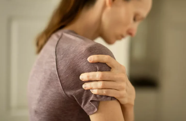 Woman in gray t-shirt clutching her shoulder in pain, representing chronic pain management during opioid addiction recovery