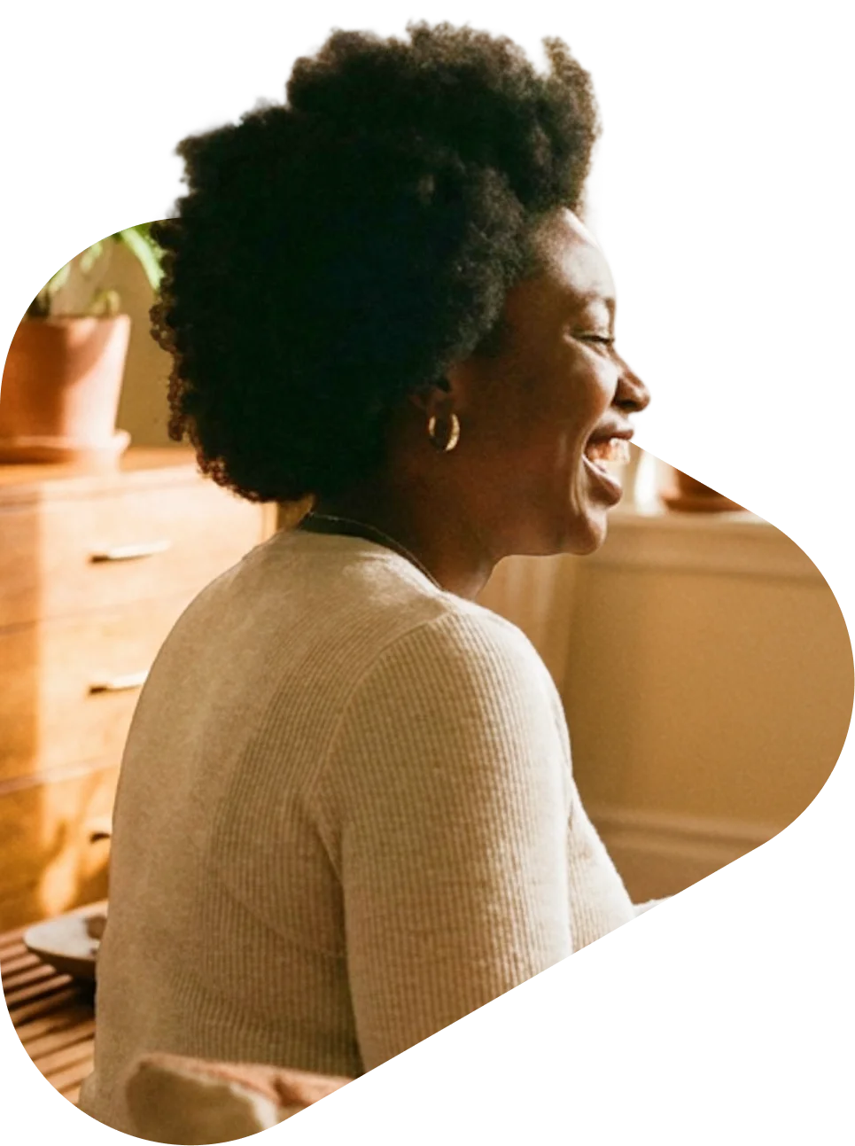 A Black woman laughing joyfully in a warm, cozy home setting wearing a beige sweater and hoop earrings.