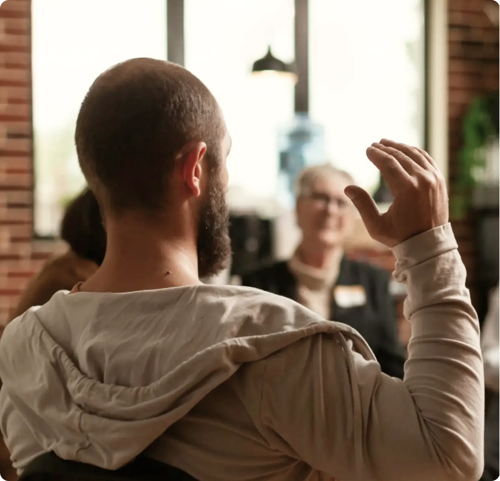 Bearded man seen from behind gesturing during a conversation with a smiling older woman in a warmly lit indoor setting