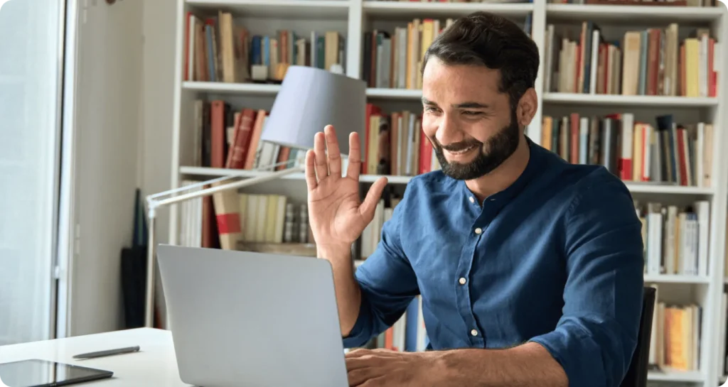 Smiling man waving during a video call on a laptop at a home desk with bookshelves in the background
