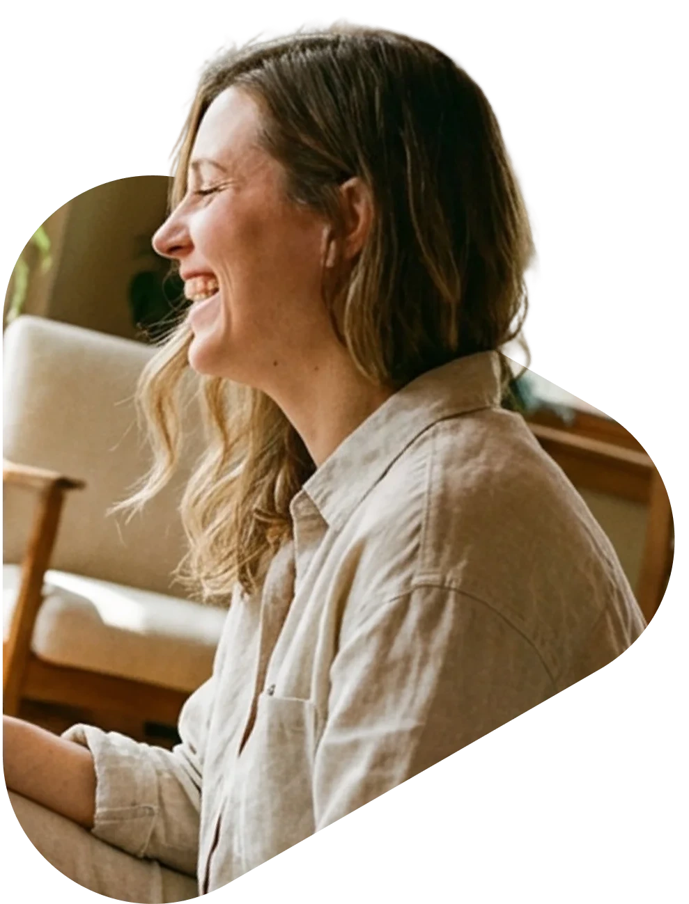 A woman with shoulder-length hair laughing joyfully while seated indoors, wearing a casual beige linen shirt.
