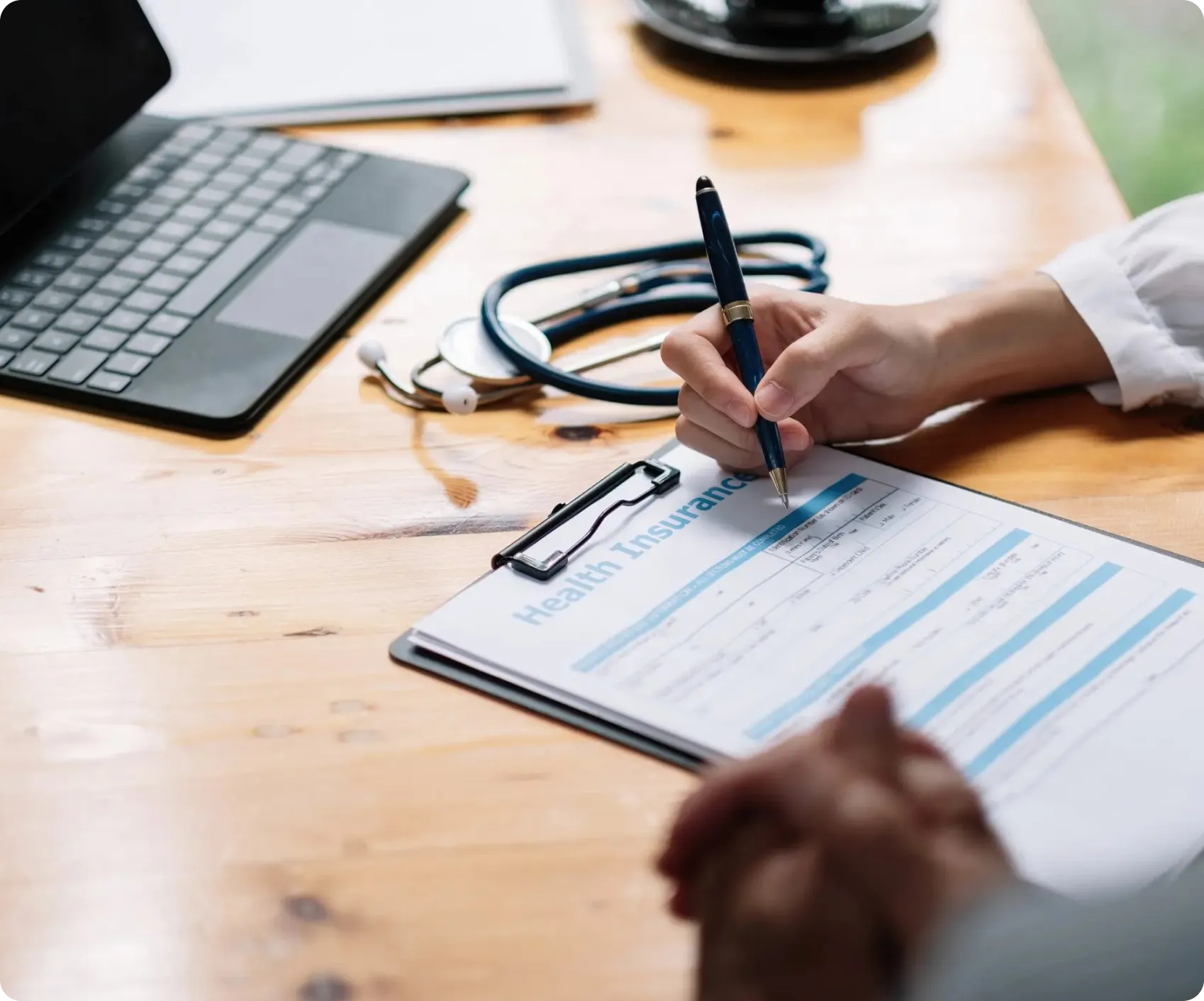Doctor filling out a health insurance form on clipboard with stethoscope and laptop on wooden desk