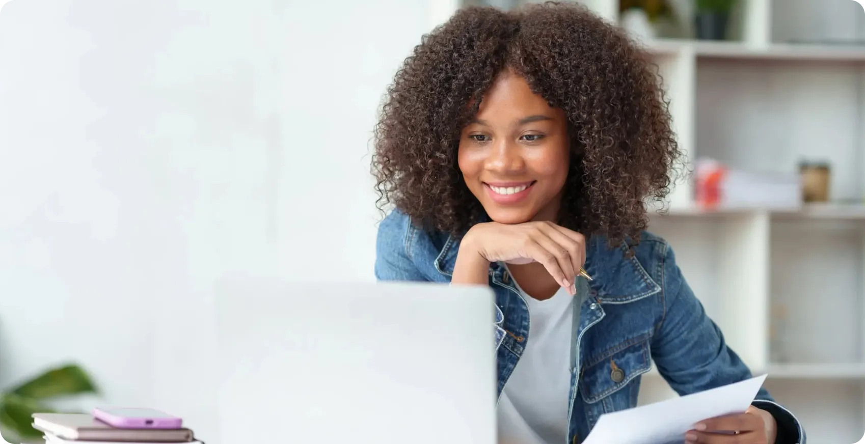 Smiling young woman with curly hair in denim jacket working on laptop, holding pen and document at desk