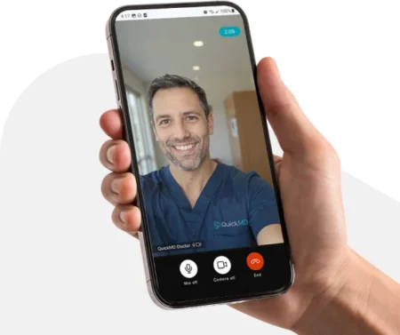 Hand holding smartphone showing a QuickMD doctor video call consultation with a smiling male physician in scrubs