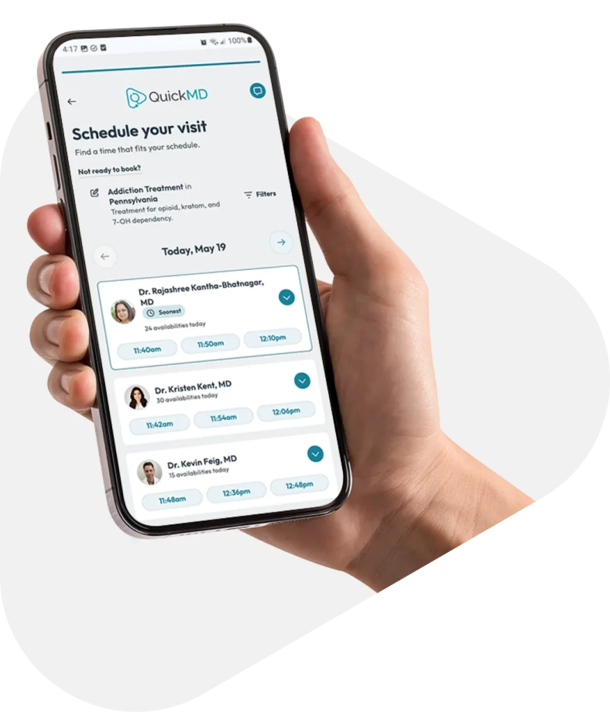 Hand holding a smartphone displaying the QuickMD app's "Schedule your visit" screen with available doctors and time slots.