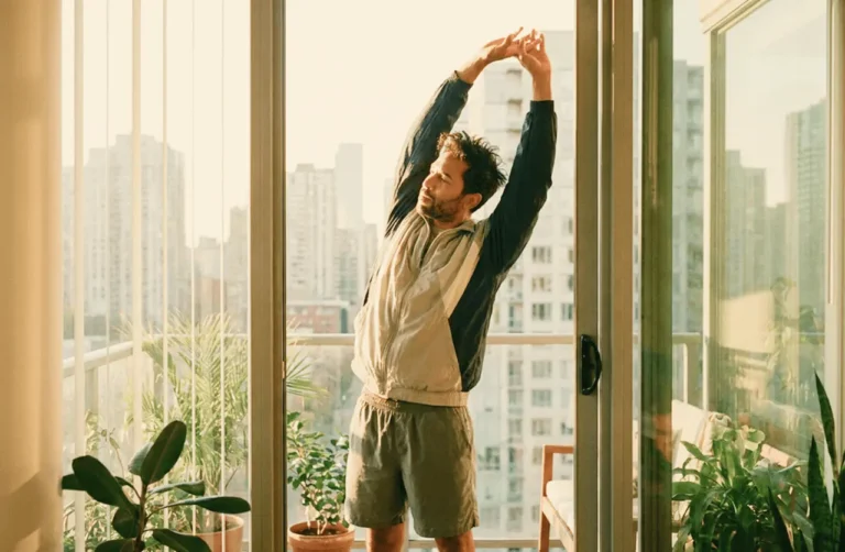 Man stretching arms overhead by a sunlit balcony door with city skyline view, surrounded by indoor plants, morning routine