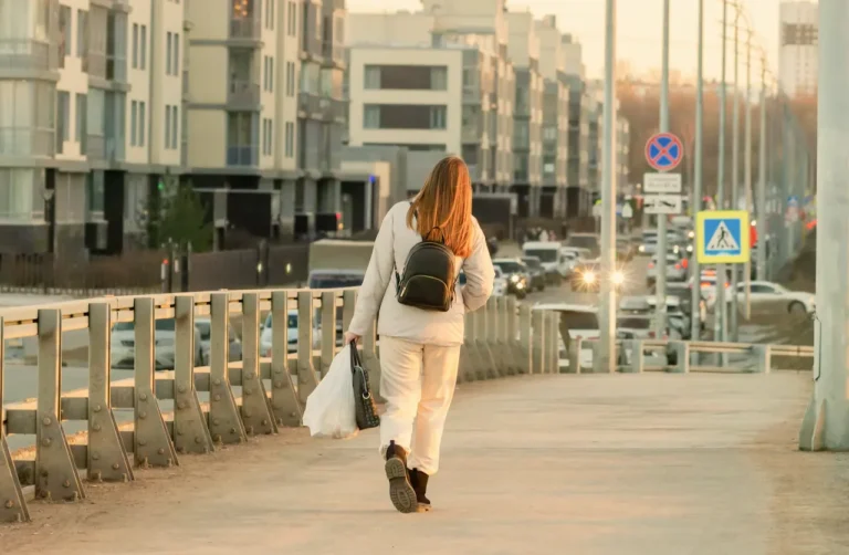 Woman walking alone on urban sidewalk with backpack, symbolizing loneliness in recovery from addiction.