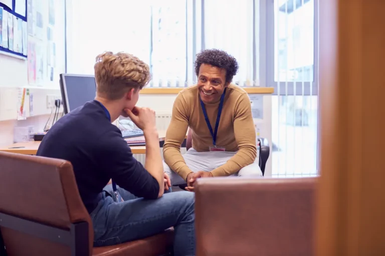 A smiling mental health counselor with a lanyard engages in a supportive one-on-one session with a young male patient in an office.