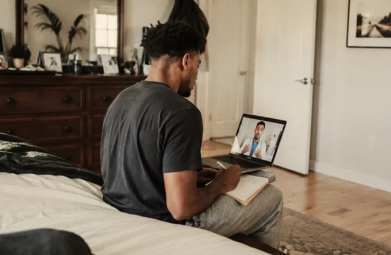 man-sitting-on-bed-taking-notes-during-a-virtual-telehealth-consultation-about-7-oh-with-a-doctor-on-laptop