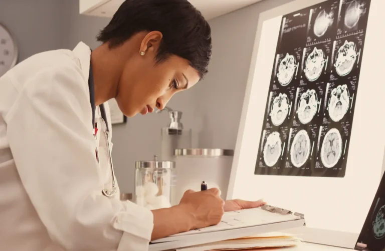 doctor-in-white-lab-coat-reviewing-brain-mri-scans-on-lightbox-while-taking-notes-on-clipboard-in-medical-office