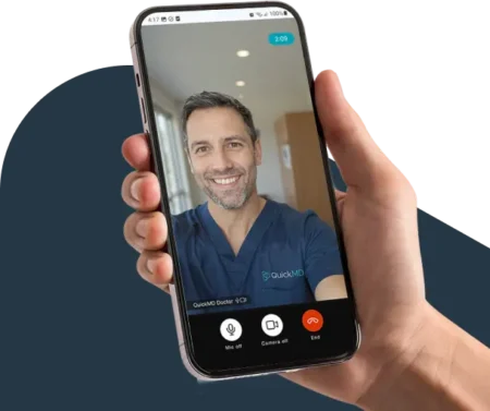 Hand holding a smartphone during a QuickMD telehealth video call with a smiling doctor in blue scrubs