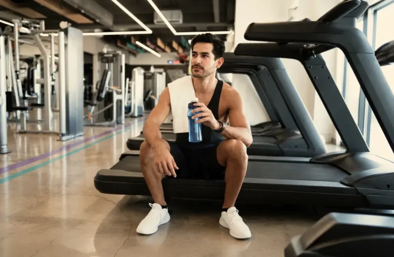 Man resting on a treadmill at the gym, holding a water bottle with a towel around his neck after working out on Suboxone