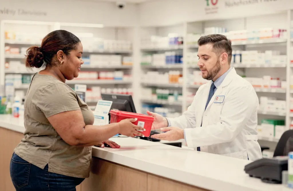 A patient hands a red sharps disposal container to a pharmacist at the pharmacy counter for safe needle disposal after GLP-1 injections